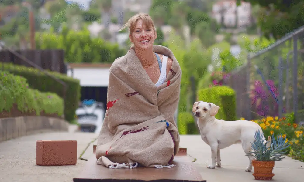 Bild von lachender Frau auf brauner Yogamatte mit Yogadecke hund in weiss rechts mit Baeumen im Hintergrund bei Hauseinfahrt