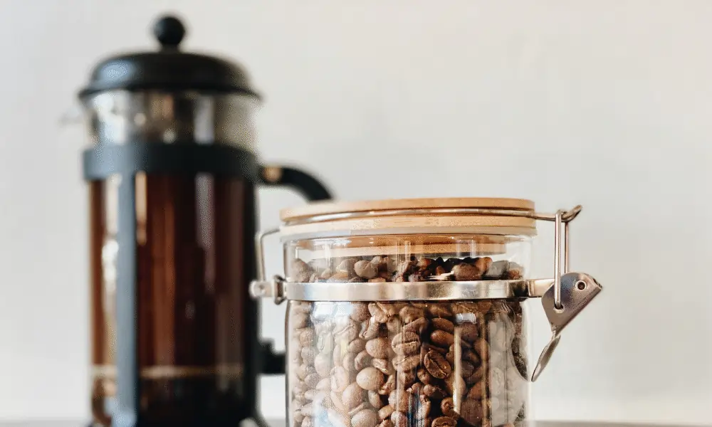 Bild von einer French Press im Hintergrund - Vordergrund Glas mit vollen Kaffeebohnen geschlossen